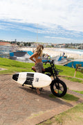 Coogee Bikes Explorer in brown carrying a surfboard on the surf rack above Bondi